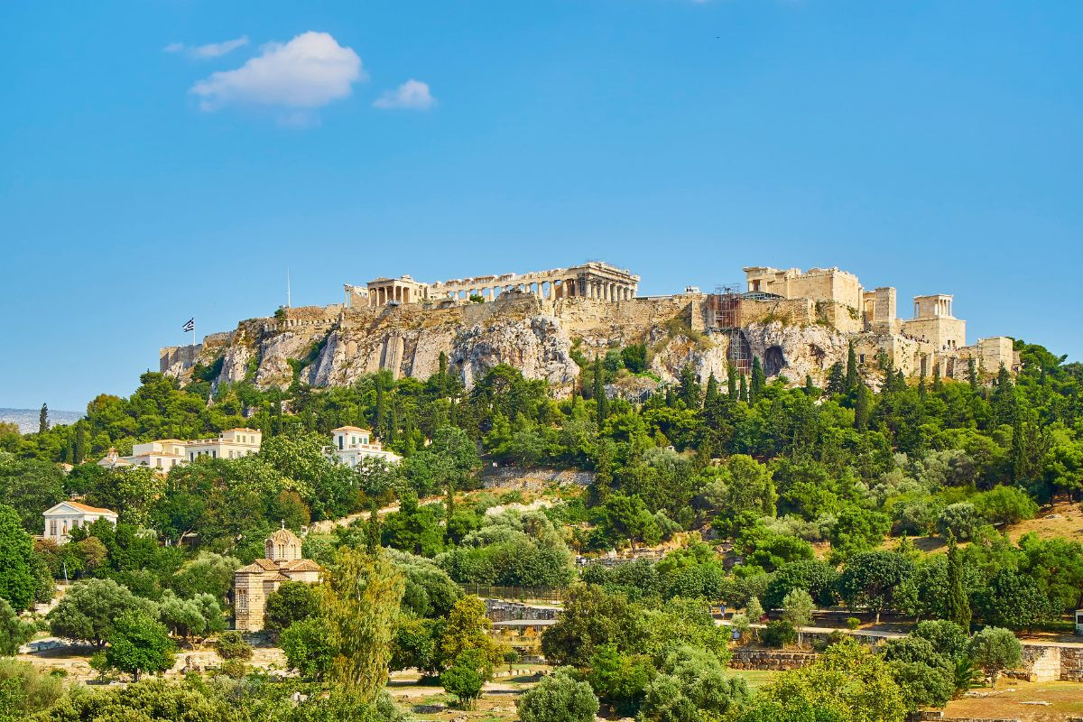 The image shows the Acropolis of Athens, a historical site situated on a rocky hill, featuring ancient Greek structures including the Parthenon. The foreground consists of lush greenery and scattered buildings, with a clear blue sky above.